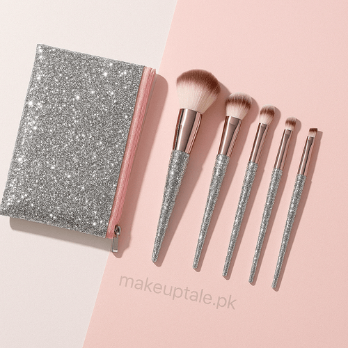 A set of five makeup brushes with glitter handles, displayed alongside a sparkling silver and pink zip pouch on a two-tone background.